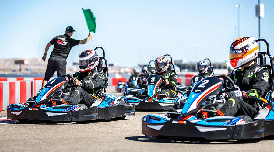 Racers in colorful go-karts speed around a sunny outdoor track. The lead driver intensely focuses ahead, conveying excitement and competition.